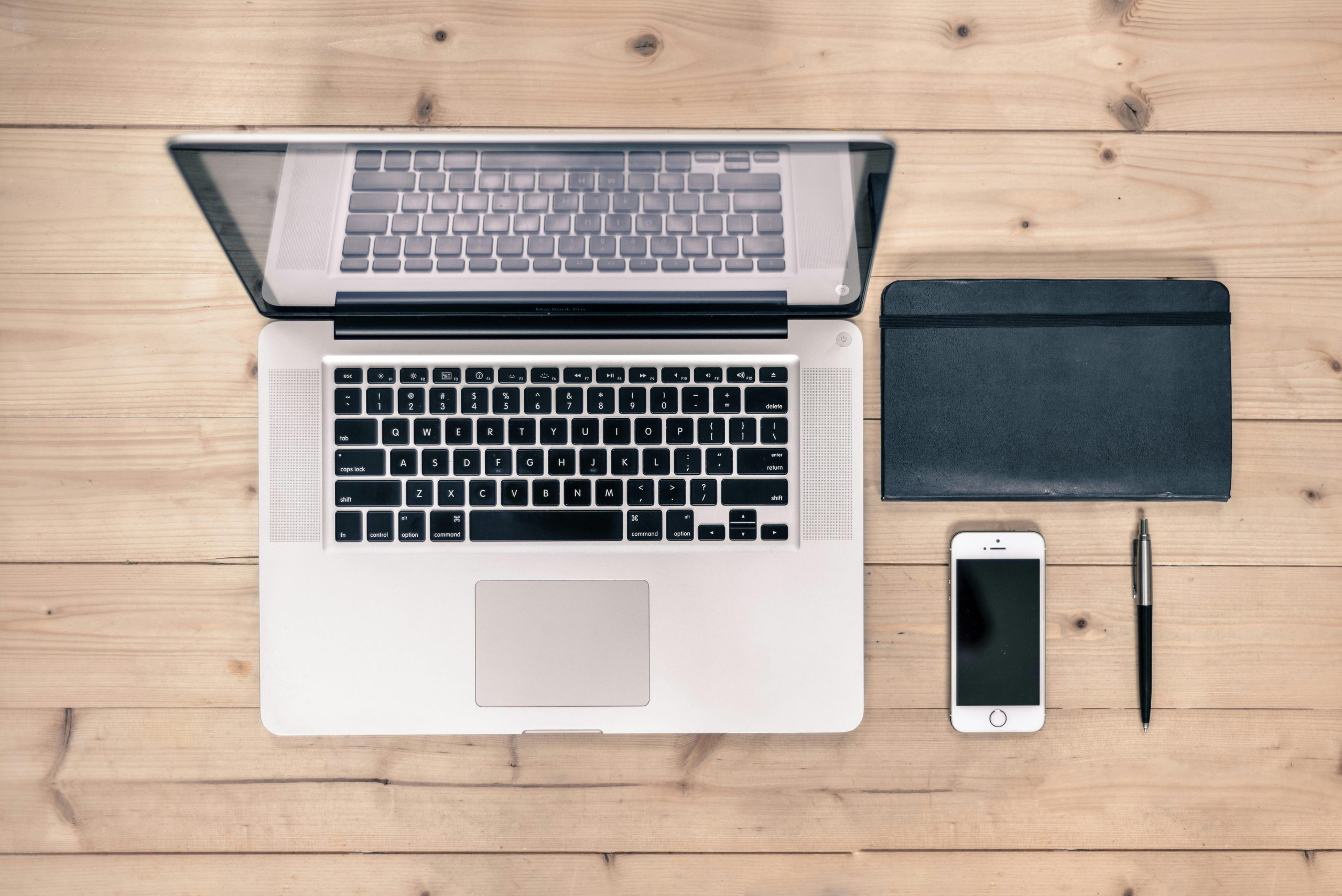 Image of a laptop, planner, cell phone and pen on a desk showing a neat, minimalist, organized space with no clutter.